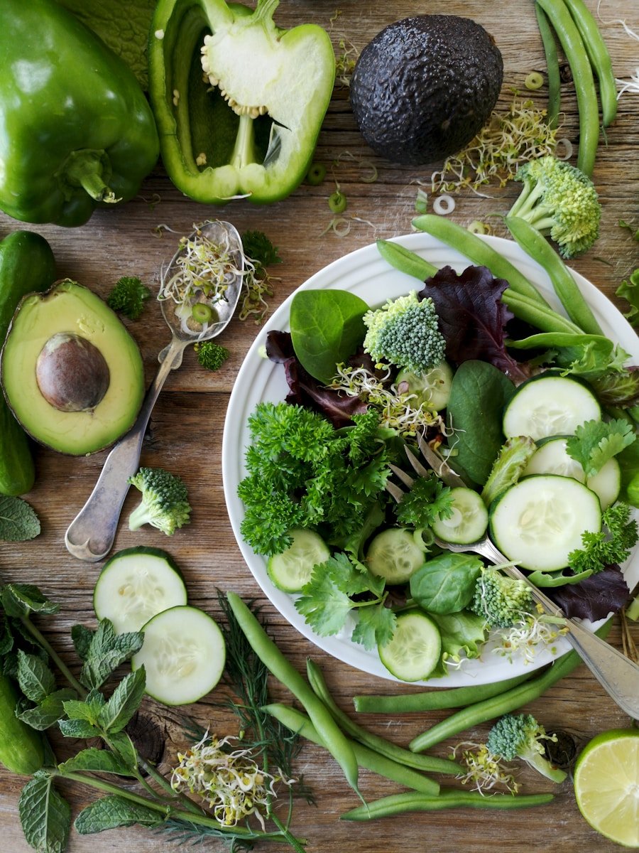 Keto Diet sliced broccoli and cucumber on plate with gray stainless steel fork near green bell pepper, snowpea, and avocado fruit